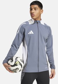 Grijze sportjasje met witte accenten, ritssluiting en zijpaneel-detailing. Model houdt een zwart-wit voetbalschoen vast. Zwarte broek.