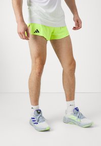 Neon gule sportsshorts med sort logo, parret med lysegrå løbesko med blå og grønne accenter, og hvide sokker.