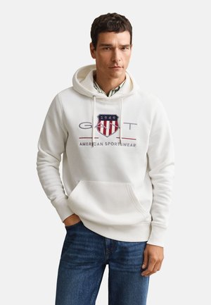 Hombre con sudadera blanca con capucha con el logo "GANT 1949 American Sportswear", vaqueros azules, de pie con una mano en el bolsillo.