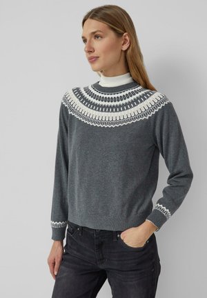 Frau mit langen blonden Haaren, die einen grauen Pullover mit weißen geometrischen Mustern und schwarze Jeans trägt, steht mit einer Hand in der Tasche.