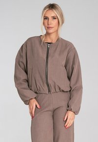 Brązowa kurtka zapinana na zamek z okrągłym dekoltem, bufiastymi rękawami i marszczoną talią. Wykonana z teksturowanej tkaniny z bocznymi kieszeniami.