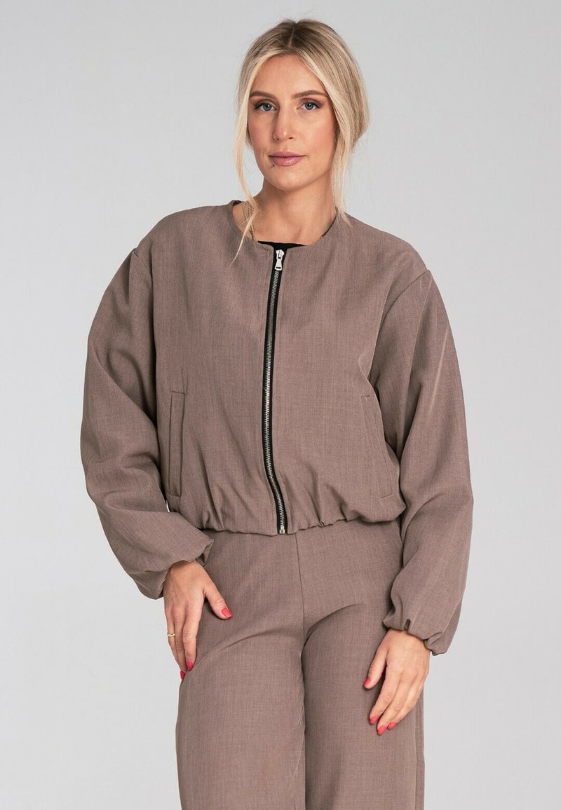 Brązowa kurtka zapinana na zamek z okrągłym dekoltem, bufiastymi rękawami i marszczoną talią. Wykonana z teksturowanej tkaniny z bocznymi kieszeniami.