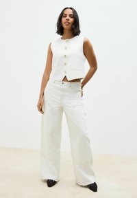Hvid ærmeløs cropped vest med guldknapper, parret med brede hvide jeans med lommer og en frynset kant. Sorte spidse sko.