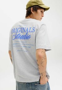 Mlad človek s tetovažami, oblečen v sivo majico z modrim napisom "ORIGINALS Studio", olivno zeleno kapo in kavbojke, vidno od zadaj.