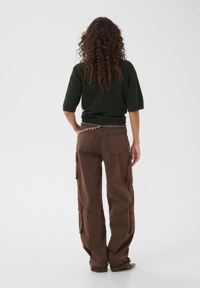 Personne aux longs cheveux bouclés, vue de dos, portant un haut à manches courtes foncé, un pantalon cargo marron et une ceinture chaîne argentée.