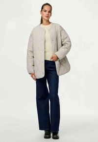 Vaaleanharmaa tikattu takki, jossa on pyöreä pääntie ja nappikiinnitys, päällä vaalea neule ja tummansiniset leveälahkeiset farkut. Mustat saappaat.