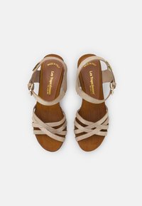 Beige suede sandals featuring crisscross straps and ankle buckle, set on a brown wood sole. Made in Italy.