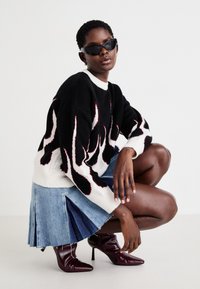 Μαύρο και άσπρο πουλόβερ πλεκτό με σχέδιο φλόγας, τζιν φούστα με πιέτες, και μπορντό γόβες με λοξό τακούνι. Φοράει μαύρα γωνιώδη γυαλιά ηλίου.