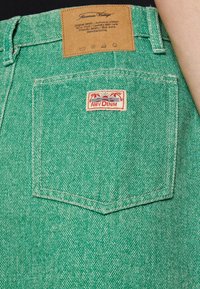 Gröna denimshorts med en texturerad yta, försedda med en läderetikett och en rödvit logotypapplikation på bakfickan.