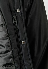 Black padded jacket detail featuring a zippered pocket, snap button, and smooth texture. The inner lining shows quilted stitching.