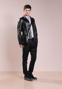 Giacca bomber in pelle nera con colletto a righe, camicia button-up di colore viola chiaro, pantaloni neri e scarpe nere eleganti, mentre tiene una piccola borsa nera.