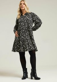 Czarna sukienka w kwiatowy wzór z długimi rękawami, wykończeniem na guziki, wielowarstwowym fasonem i kieszeniami, zestawiona z czarnymi botkami za kostkę. Gładka tekstura.