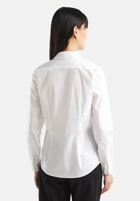 Camicia bianca aderente con colletto classico, cucitura posteriore e maniche lunghe. Tessuto di cotone liscio, orlo curvato.