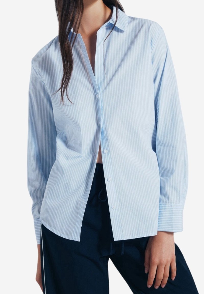 Femme portant une chemise bleu clair à rayures verticales, boutonnée, à manches longues et un pantalon foncé sur fond blanc.