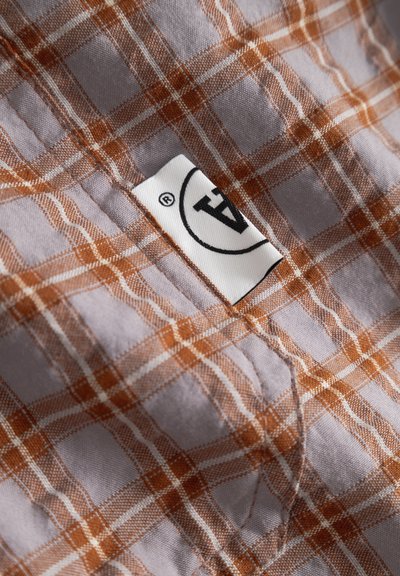 Tissu violet clair avec un motif à carreaux orange et blanc, montrant une poche cousue et une étiquette de vêtement blanche partiellement visible avec une impression noire.