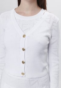 Witte gebreide cardigan met een v-hals, voorzien van gouden decoratieve knopen en een textuurpatroon van kleine geometrische vormen.