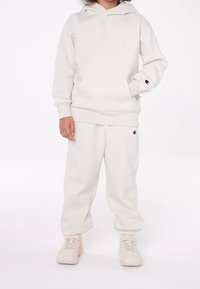 Barn i krämfärgad Champion-hoodie och sweatpants med matchande krämfärgade sneakers, som står mot en vit bakgrund.
