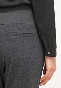 Harmaat villahousut, joissa on takatasku ja sileä pinta. Musta paita, jossa on napitettu hihansuu, näkyy osittain vasemmalla.