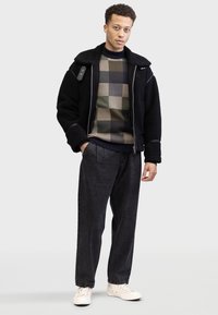 Veste en shearling noire, pull à carreaux vert et beige, pantalon gris foncé, baskets blanches, avec des coutures détaillées et des fermetures éclair.