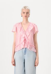 Blusa rosa con rouches, scollatura a V e maniche corte, decorata con volant lungo il davanti e l'orlo, abbinata a pantaloni in denim azzurro chiaro.