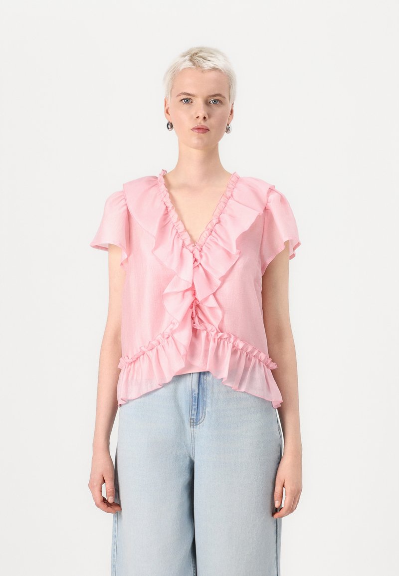 Blusa rosa con rouches, scollatura a V e maniche corte, decorata con volant lungo il davanti e l'orlo, abbinata a pantaloni in denim azzurro chiaro.