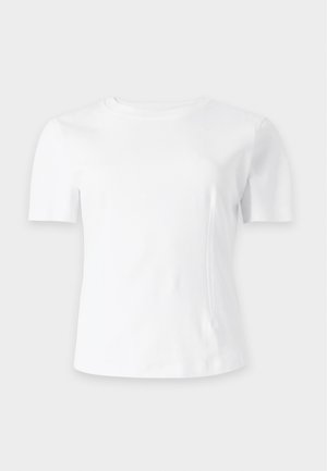Femme aux cheveux noirs raides portant un haut blanc à manches courtes et un pantalon large noir ample, debout les mains dans les poches sur fond blanc.