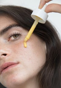 Droppflaska doserar ett gult serum på huden. Flaskan har en vit kork och en smal, genomskinlig droppe.