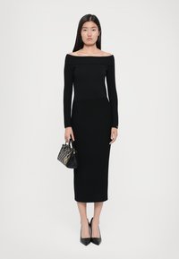Musta ribbed midi-mekko, jossa on olkaimeton kaula-aukko, pitkät hihat ja vartalonmyötäinen siluetti. Yhdistettynä teräväkärkisiin mustiin korkokenkiin ja pieneen käsilaukkuun.