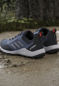 Paire de chaussures de randonnée Adidas Terrex bleues et noires sur un sol en gravier humide près des racines d’arbres et de la végétation.