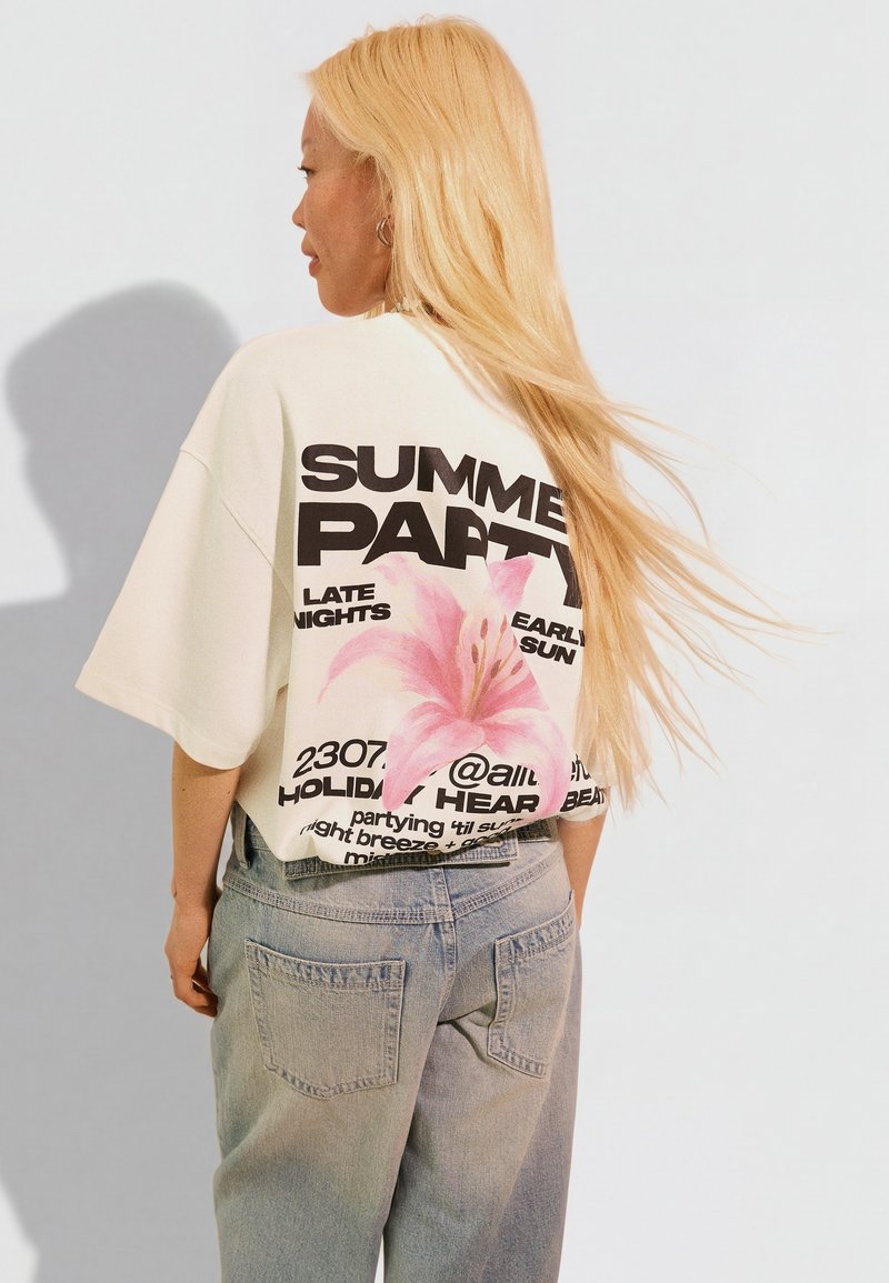 Femme aux longs cheveux blonds portant un t-shirt court blanc oversize avec le texte « SUMMER PARTY » et un motif de fleur rose, assorti à un jean bleu clair.