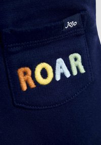 A tengerészkék anyagon egy hátsó zseb található, amelyen több színben - narancssárga, sárga, fehér és zöld - hímzett "ROAR" felirat látható.