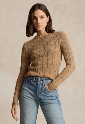 Woman with shoulder-length dark hair wearing a camel-colored cable knit sweater and light blue high-waisted jeans, posing with one hand in pocket.