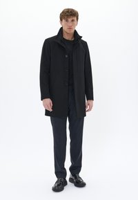 Manteau en laine noir avec un col montant, cinq boutons et deux poches latérales. Porté sur une chemise noire et un pantalon à motifs sombres, accompagné de chaussures noires brillantes.