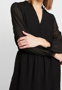 Schwarzes Kleid mit tiefem V-Ausschnitt, Knopfdetails, durchsichtigen langen Ärmeln und elastischen Bündchen. Flauschige Silhouette mit strukturiertem Stoff.