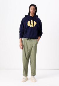 Hanorac bleumarin cu logo-ul galben "GAP", buzunar tip cangur; asociat cu pantaloni tapered verzi deschis și adidași bej. O țesătură texturată și o croială lejeră.