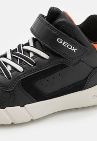 Zapatilla negra de cuero con acentos de ante gris, cordones blancos, tira de Velcro y logotipo de Geox. Cuenta con detalles perforados y una suela de goma blanca.