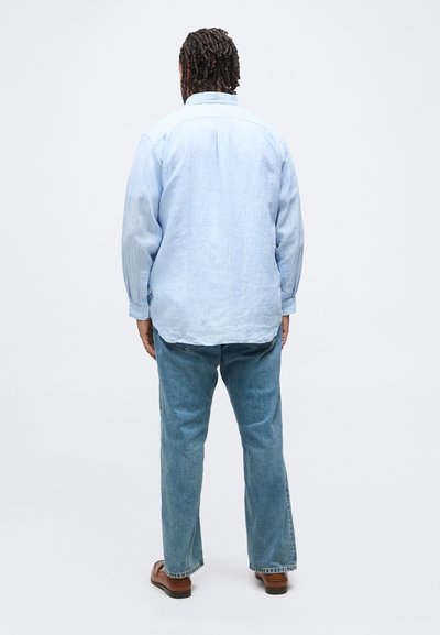 Άνδρας με πλεξούδες στα μαλλιά, φορώντας ένα ανοιχτό μπλε ριγέ πουκάμισο, μπλε τζιν και καφέ παπούτσια, στέκεται γυρισμένος προς τα πίσω μπροστά σε ένα απλό λευκό φόντο.