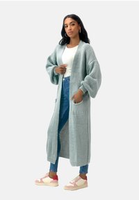 Stickad lång kofta i ljus mintgrön, med översized ärmar, två framfickor och en avslappnad passform, i kombination med blå jeans och sneakers.