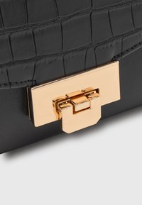 Coin d'un sac à main en cuir noir texturé avec une boucle rectangulaire dorée brillante sur un fond pâle.