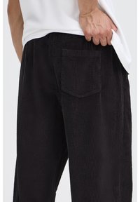 Pantalones de pana negros con estrías verticales, con un bolsillo trasero y un ajuste relajado, combinados con una camiseta blanca lisa.