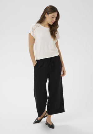 Žena v bielom top s čipkovanými ramenami a voľnými čiernymi nohavicami, stojaca s rukami v vreckách, pozerajúca sa dole.