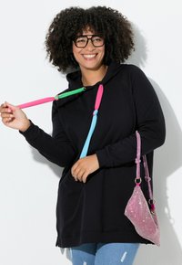 Donna sorridente con capelli ricci e occhiali, che indossa un hoodie nero con lacci colorati e tiene una borsa di strass rosa sotto il braccio.