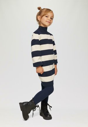 Gestreepte jurk in een crème en marineblauwe wolmix, met een hoge hals en driekwartmouwen, gecombineerd met marineblauwe leggings en zwarte veterschoenen.