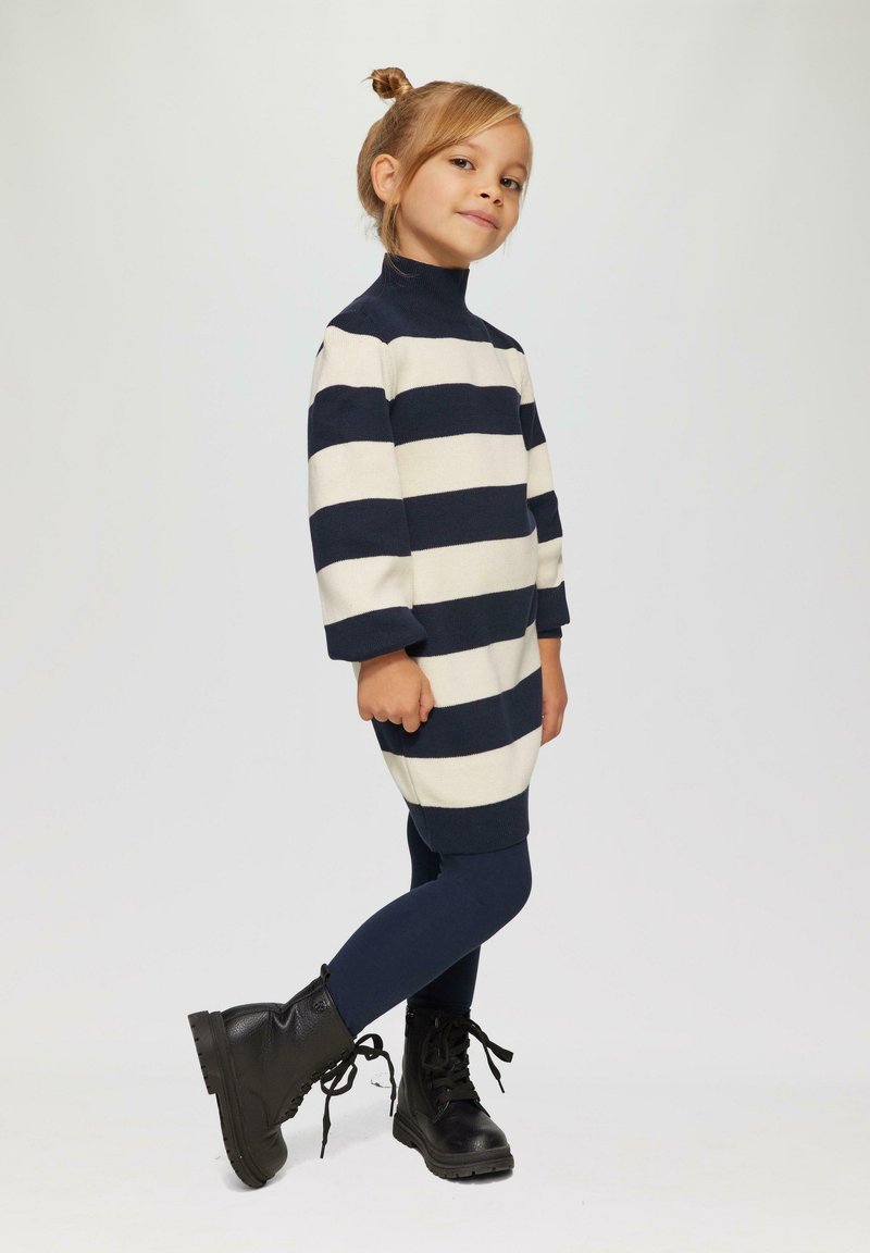 Gestreiftes Kleid aus einer cremefarbenen und marineblauen Wollmischung, mit hohem Kragen und Dreiviertelärmeln, kombiniert mit marineblauen Leggings und schwarzen Schnürstiefeln.