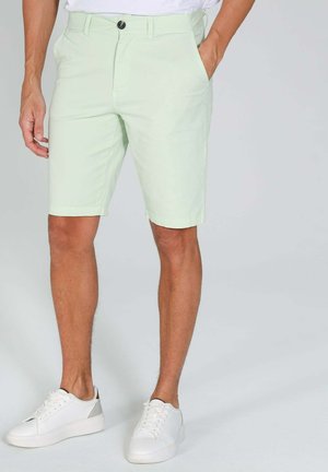 Homme portant un short vert clair jusqu'aux genoux et des baskets blanches, debout avec une main dans la poche sur un fond uni.