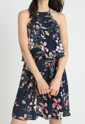 Žena v tmavě modrých bezrukávových květovaných šatech se vzory růžových, bílých a zelených květin, stojící se sepjatýma rukama před sebou.