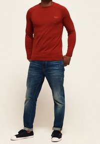 Maglione rosso a maniche lunghe in tessuto texturizzato con scollo rotondo, abbinato a jeans di denim blu con vestibilità rilassata e orli arrotolati, indossato con sneakers nere.