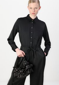 Zwarte button-up jurk met een elastische taille, lange mouwen en een borstzak; gecombineerd met een zwarte handtas met bloemmotief.