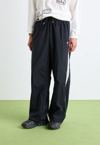 Pantalons de sport légers noirs avec une taille élastique, des panneaux latéraux blancs et un petit logo près de l'ourlet ; associés à des chaussures noires à textures multiples.
