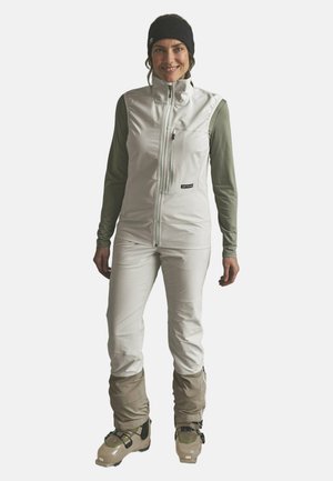 Mujer con pantalones de esquí de color claro, chaleco blanco sobre camisa de manga larga, cinta negra en la cabeza y botas de esquí beige, de pie y sonriendo.